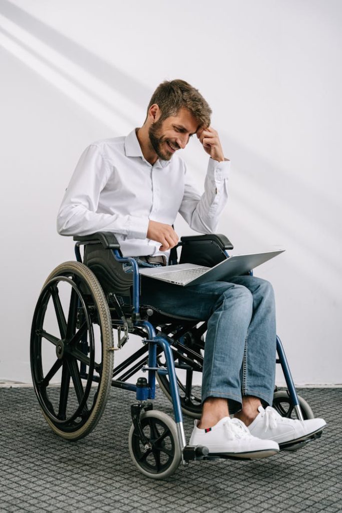 a-man-sitting-on-the-wheelchair-8127976 Man in wheelchair working on a laptop indoors, showcasing accessibility and technology.