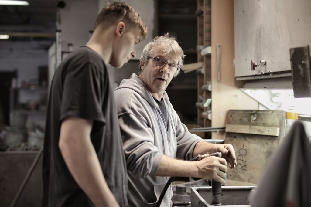 workers-handling-detail-by-pneumatic-tool-in-workshop-3846262 Side view of senior foreman in eyeglasses showing to trainee how handling detail in workshop